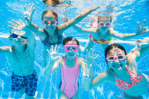 Kids under water in a swimming pool wearing googles and smiling at the camera